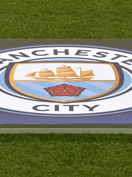 Das Bild zeigt das Logo des englischen Fußballvereins Manchester City. Es liegt im Stadion ausgebreitet auf dem Rasen.
