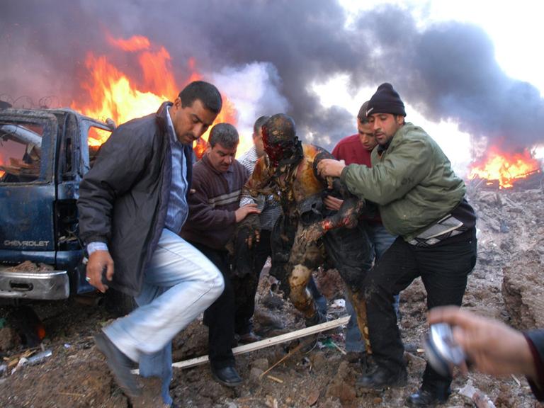 Libanesen retten einen mit Dreck beschmierten Überlebenden des Anschlags in Beirut, im Hintergrund lodern flammen.