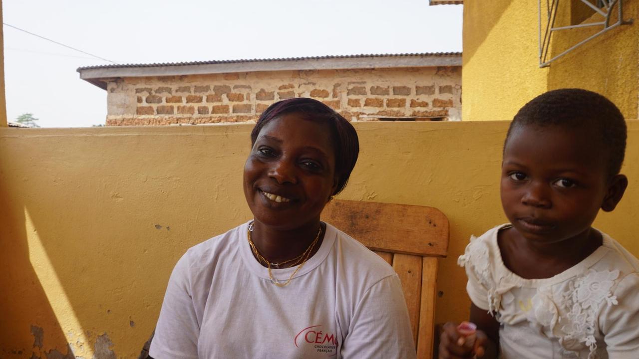 Eine Frau und ihr Kind - beide in weißen T-Shirts - sitzen auf einem Balkon und lächeln in die Kamera.