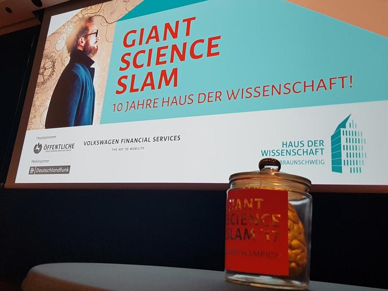 In einem großen Glas befindet sich ein goldfarbenes Hirn. Das Glas steht auf einem Tisch, im Hintergrund ist auf der Leinwand das Logo des Giant Science Slam zu sehen.