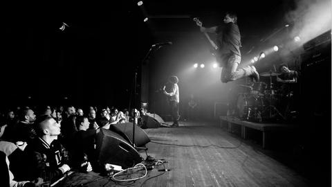 Drei Männer spielen Rockmusik auf einer Bühne, der Bassist im Bildvordergrund springt gerade in die Luft.