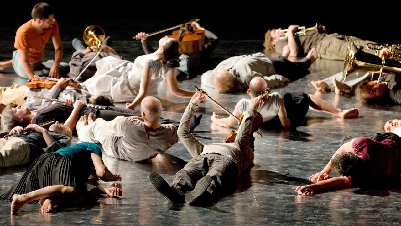 Musikerinnen und Musiker liegen mit ihren Instrumenten in den Händen auf einer Bühne.