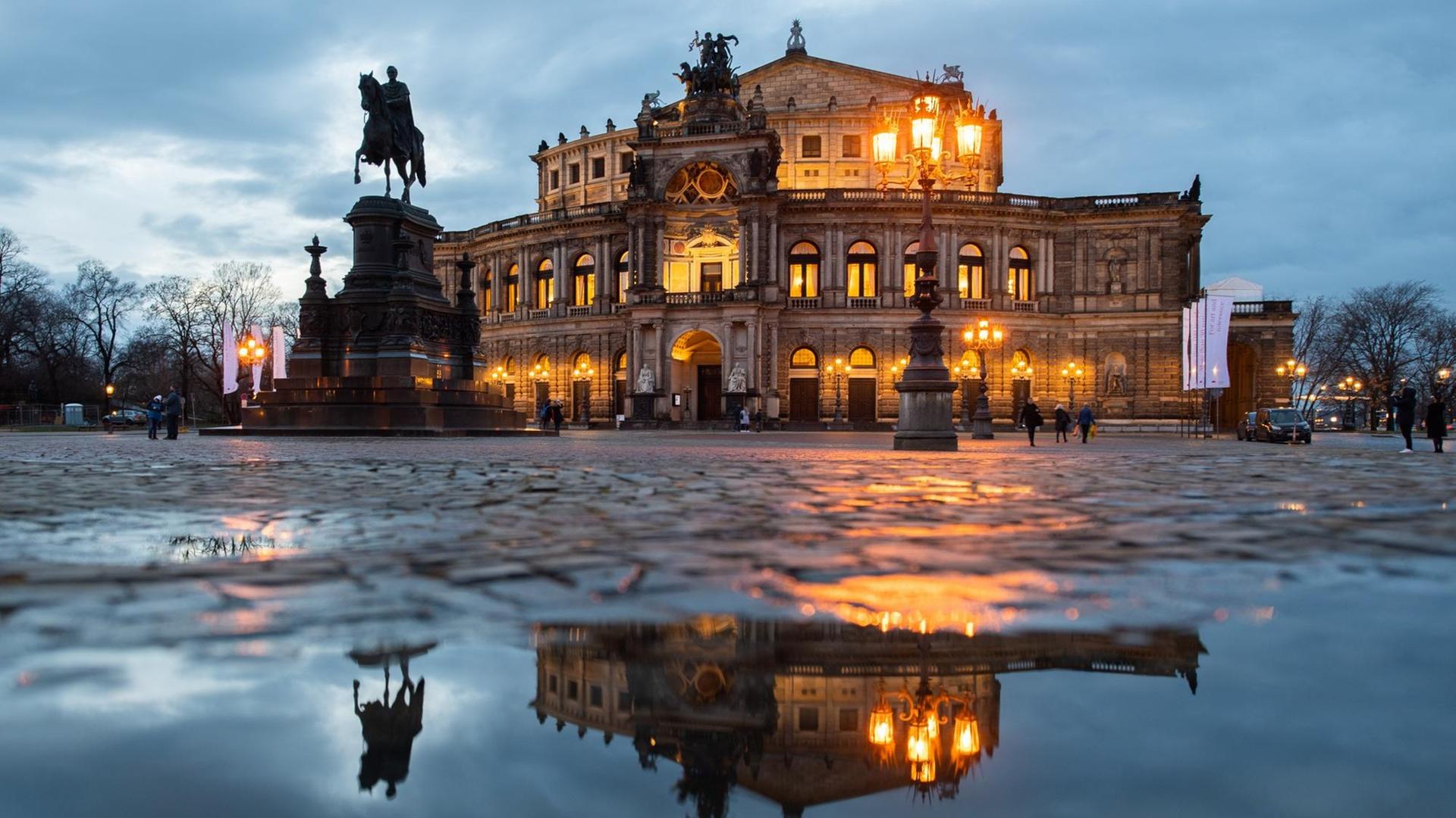 Die Semperoper und das König-Johann-Denkmal spiegeln sich am Abend in einer Pfütze auf dem Theaterplatz in Dresden.