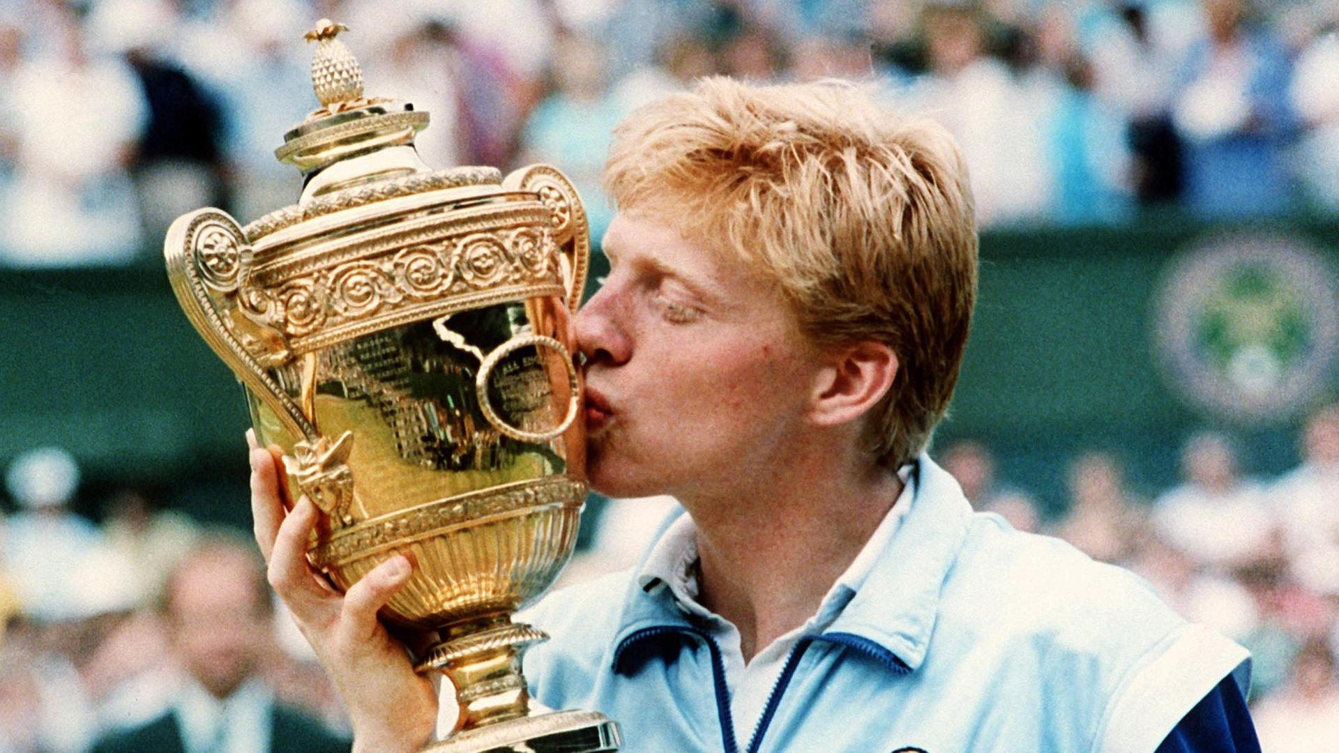 Becker in hellblau-dunkelblauen Trainigsjacke küsst den goldenen Pokal.