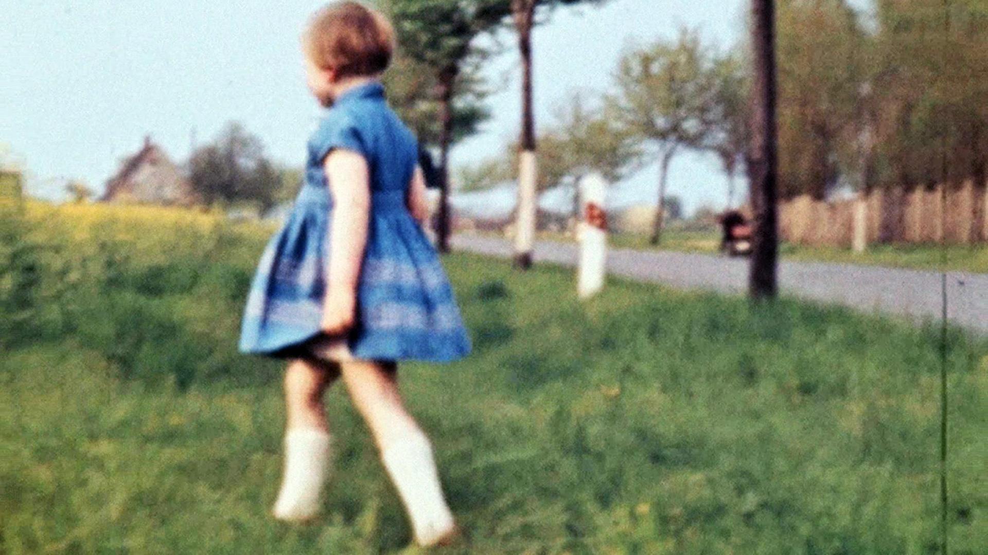 Ein Mädchen in einem Kleid läuft über eine Wiese, im Hintergrund ist eine Allee zu sehen, typisch körnige DDR-Film_Aufnahme