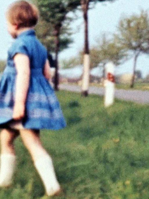 Ein Mädchen in einem Kleid läuft über eine Wiese, im Hintergrund ist eine Allee zu sehen, typisch körnige DDR-Film_Aufnahme