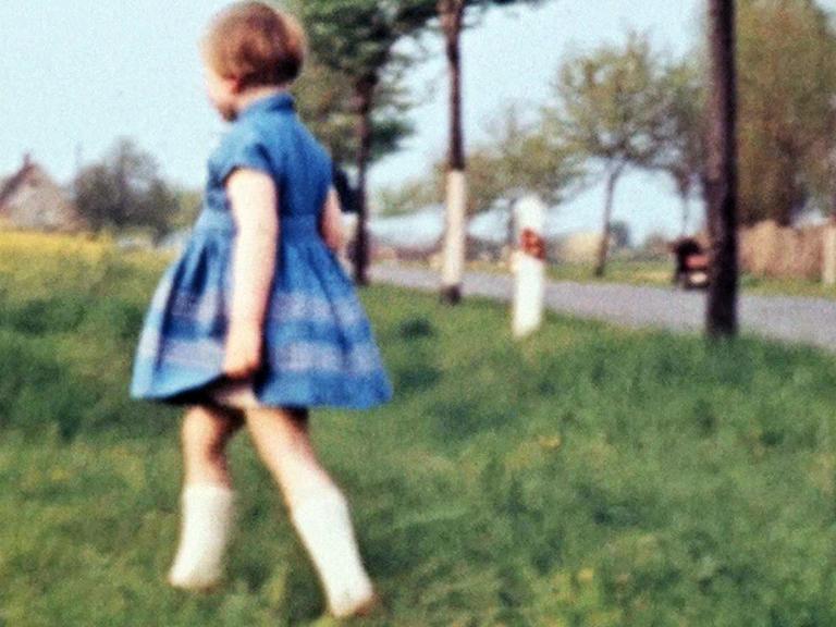 Ein Mädchen in einem Kleid läuft über eine Wiese, im Hintergrund ist eine Allee zu sehen, typisch körnige DDR-Film_Aufnahme