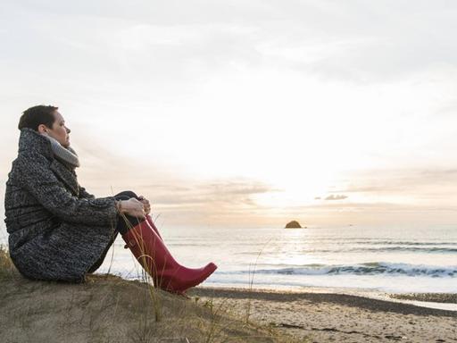 Eine Frau sitzt in Mantel und Gummistiefeln allein an einem Strand in Frankreich.