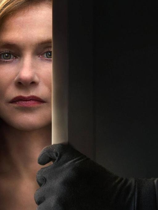 Die Schauspielerin Isabelle Anne Huppert ist durch den Spalt einer Tür zu sehen, eine Hand in schwarzem Handschuh öffnet die Tür