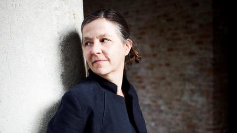 Eine Frau in dunkelblauem Jackett lehnt sich an eine weiße Wand und blickt lächelnd über die Schulter.