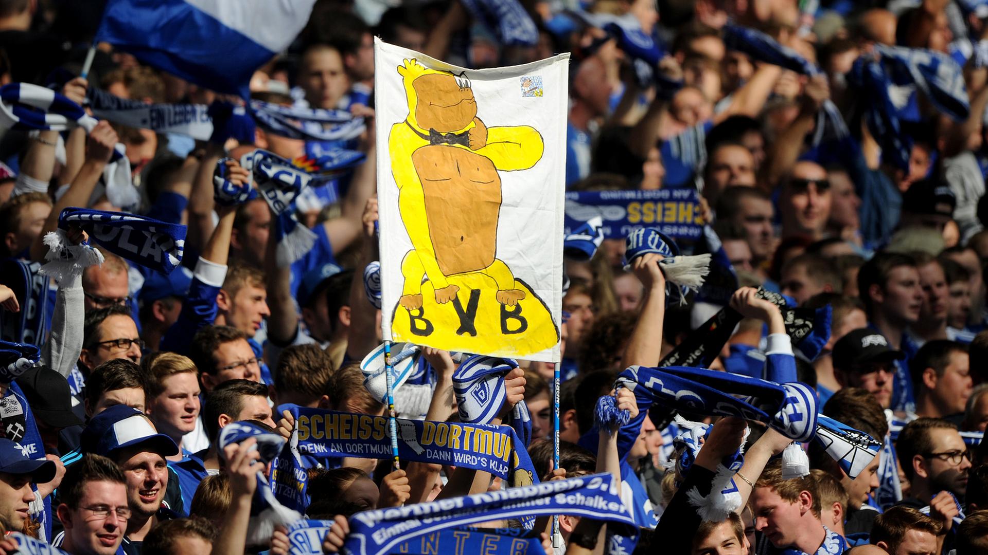 Schalke-Fans halten vor Spielbeginn ein Banner hoch, auf dem ein Affe in einem gelben Kostüm auf dem BVB-Vereinswappen sitzt.