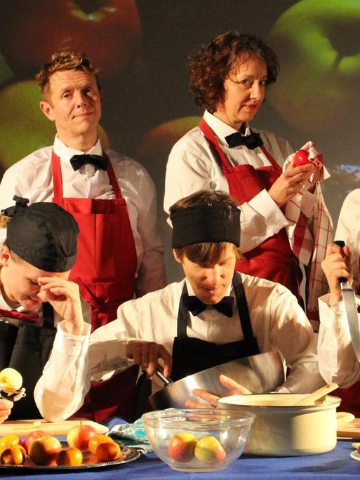 Das deutsch-britische Theaterkollektiv Gob Squad. Das Foto ist eine Gruppenaufnahme, die Bezug nimmt auf die aktuelle Inszenierung des Stücks. Die sieben Schauspieler posieren mit Kochschürzen und Kochzutaten.