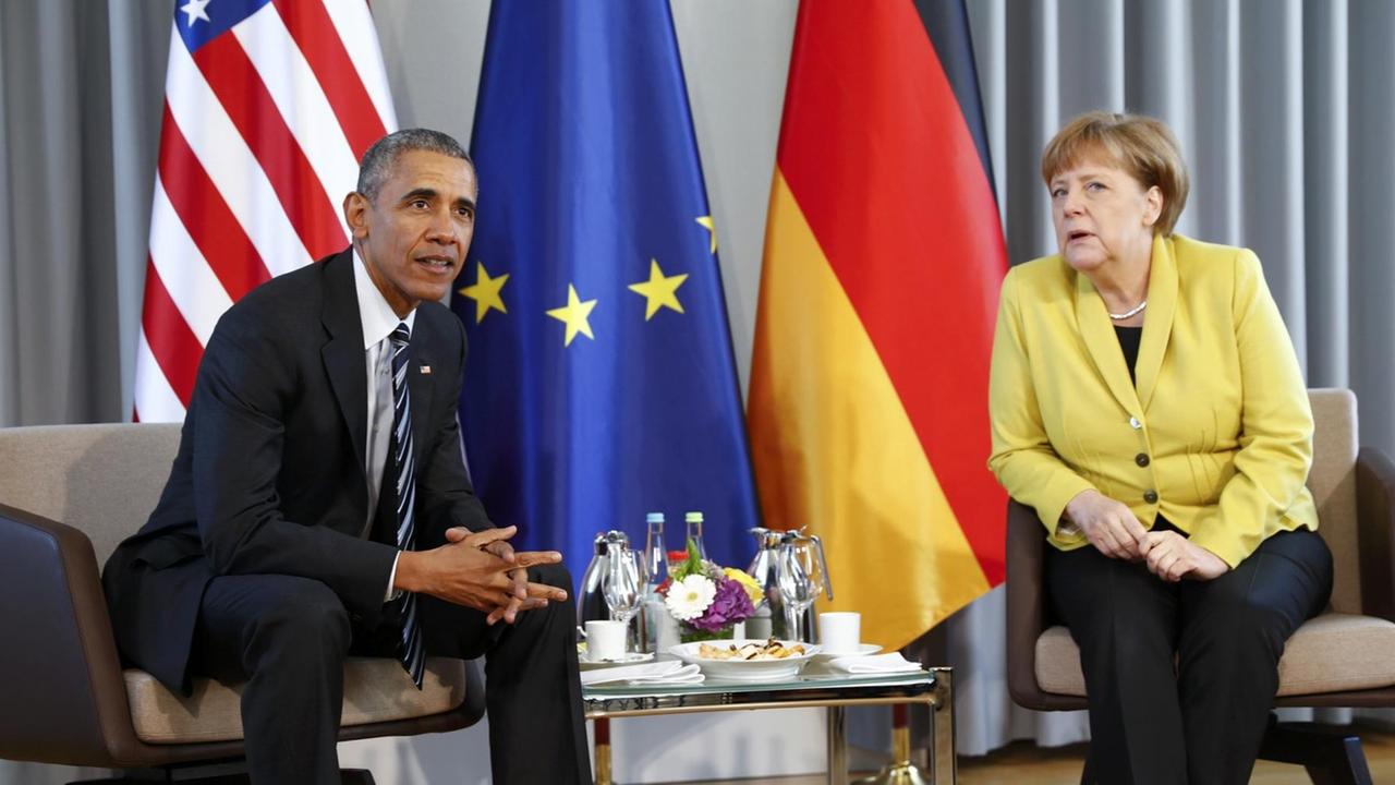 Sie sehen Barack Obama und Angela Merkel, dahinter die Fahnen der USA, der EU und Deutschlands.