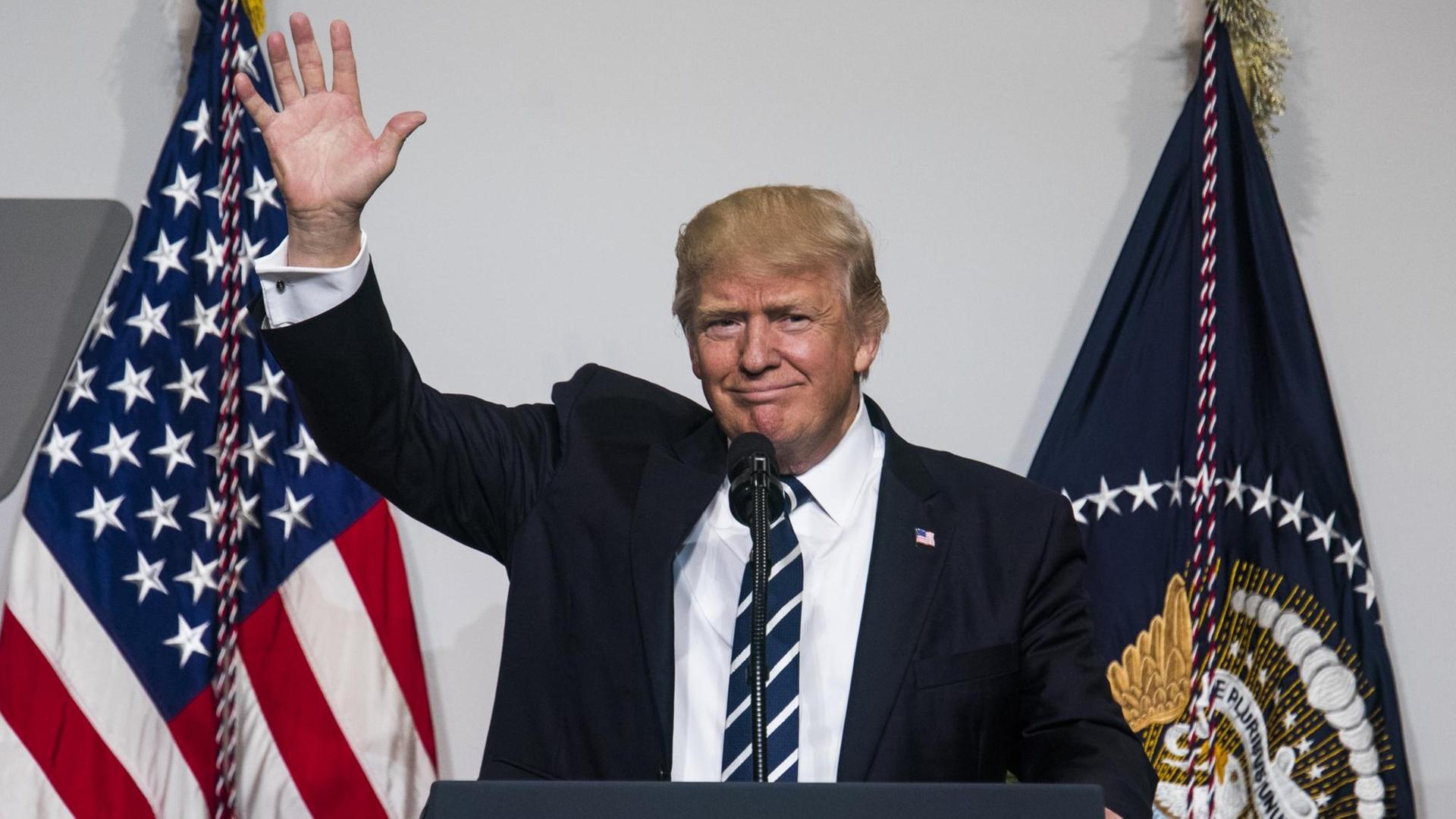 US-Präsident Donald Trump hebt seine rechte Hand zum Gruß, während er hinter einem Rednerpult und vor einer amerikanischen Flagge steht