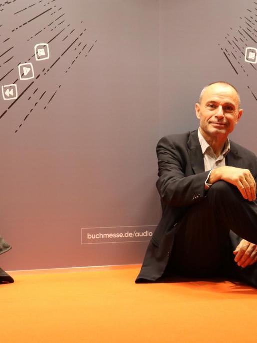Norbert Gstrein sitzt auf einem orangenen Boden und lehnt an einer Wand.