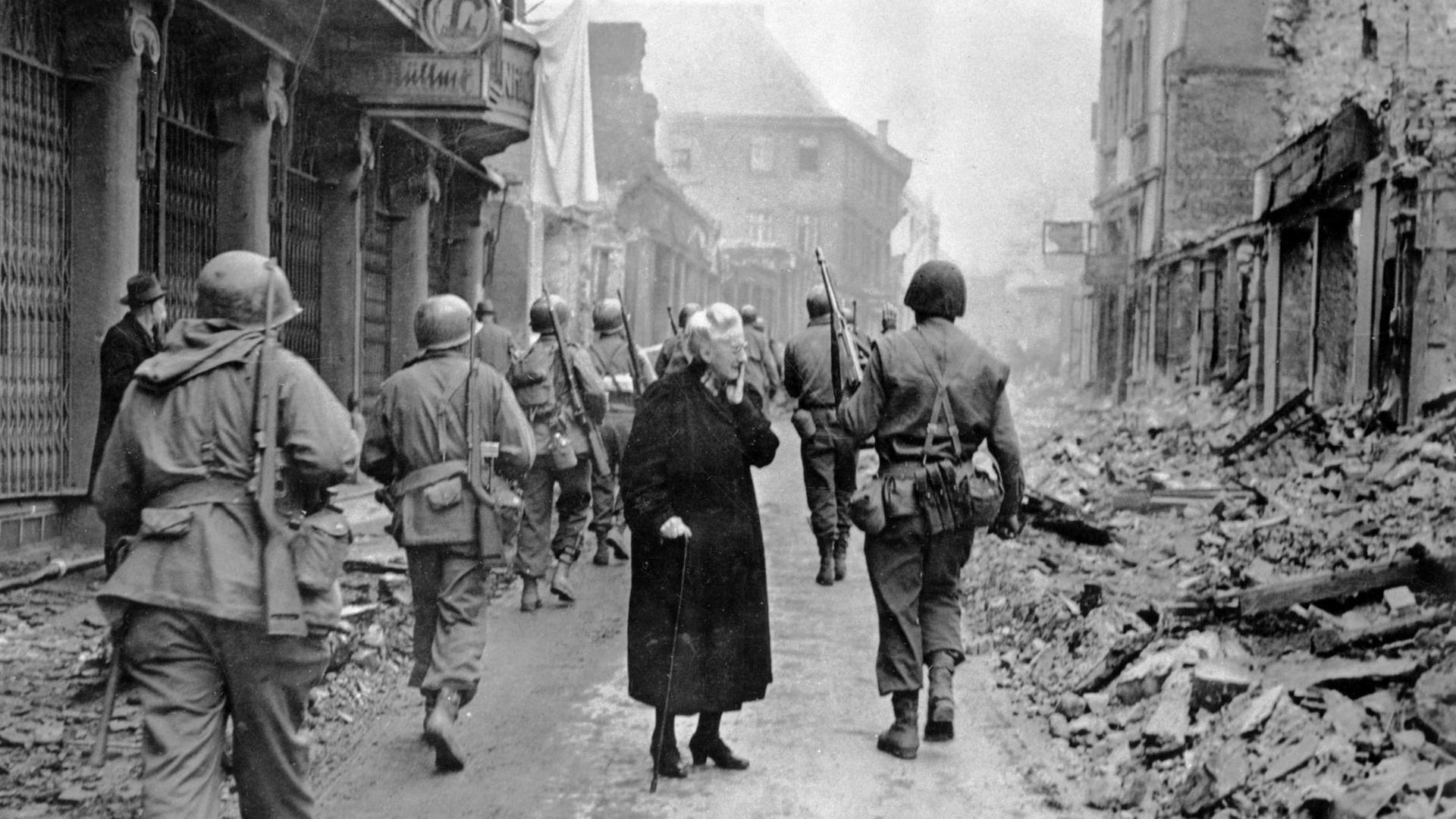 Fassungslos steht eine alte Frau in den Trümmern der Stadt Bensheim, während US-Soldaten an ihr vorbei marschieren.