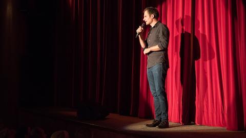 Moderator Theo Vagedes steht auf der Bühne vor einem roten Samtvorhang.