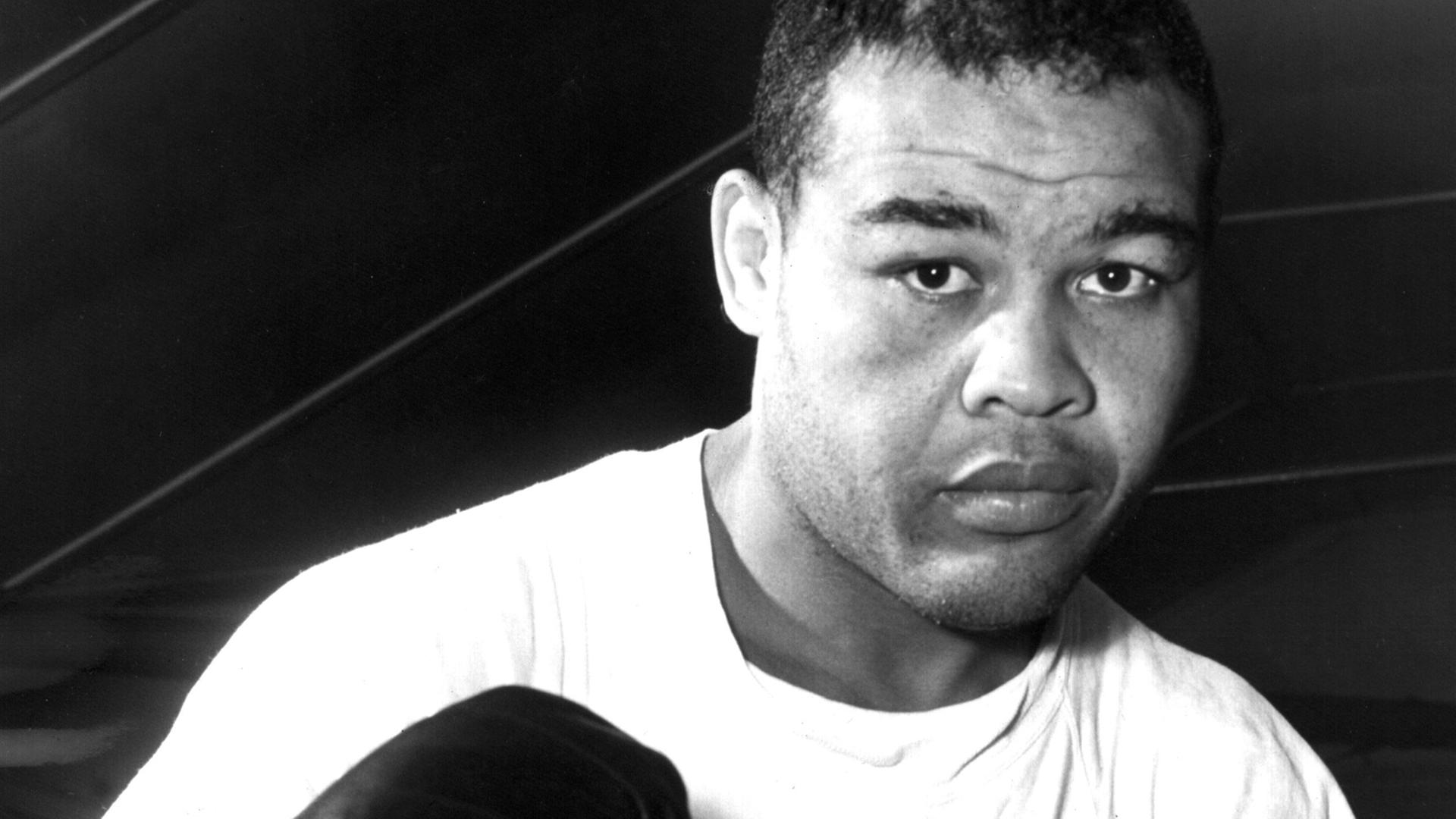 Zu sehen ist der afroamerikanische Boxer Joe Louis in einer Aufnahme von 1950. Er trägt ein weißes T-Shirt und schwarze Boxhandschuhe und präsentiert die Fäuste in typischer Boxermanier dem Betrachter.