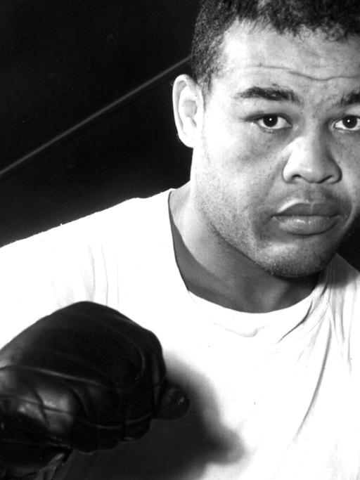 Zu sehen ist der afroamerikanische Boxer Joe Louis in einer Aufnahme von 1950. Er trägt ein weißes T-Shirt und schwarze Boxhandschuhe und präsentiert die Fäuste in typischer Boxermanier dem Betrachter.