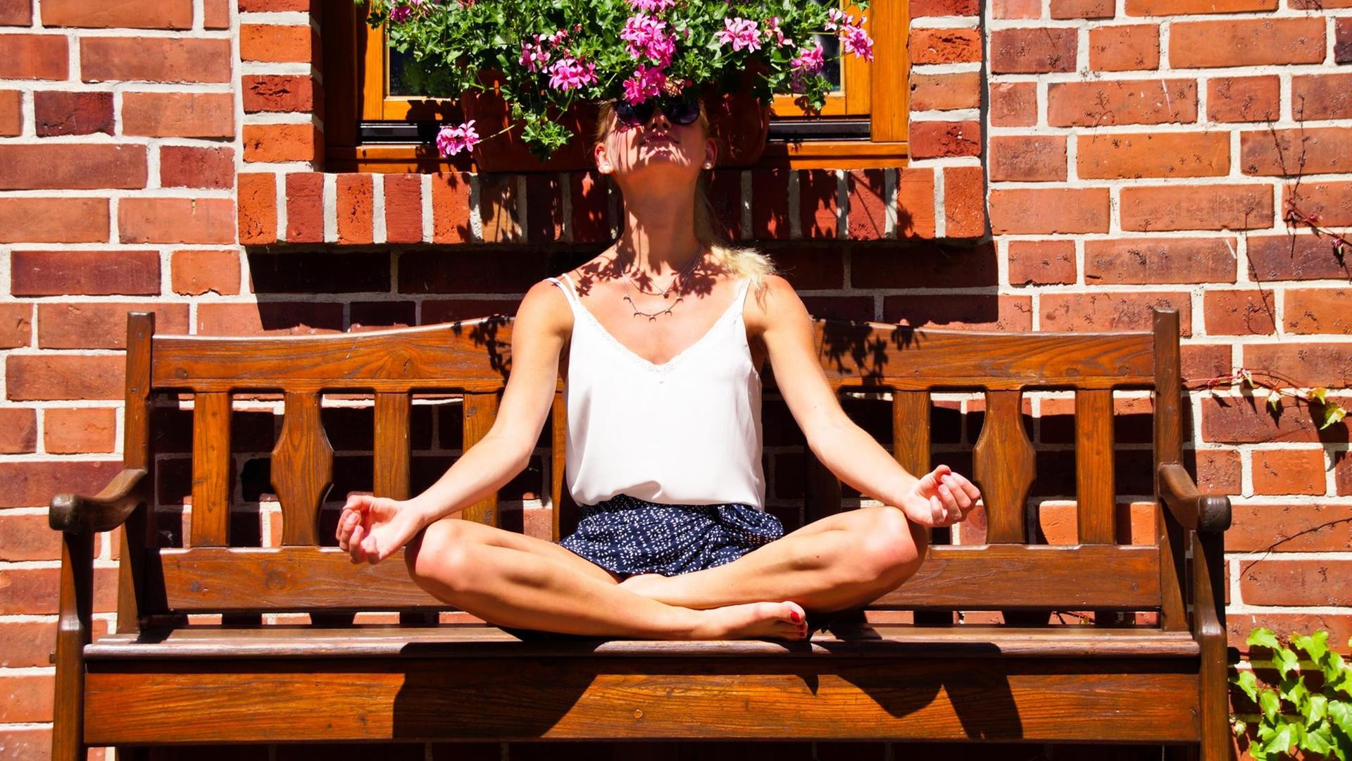 Eine junge Frau sitzt im Schneidersitzt auf einer Bank. Sieht atmet dabei den Geruch der Blumen über ihren Kopf tief ein.