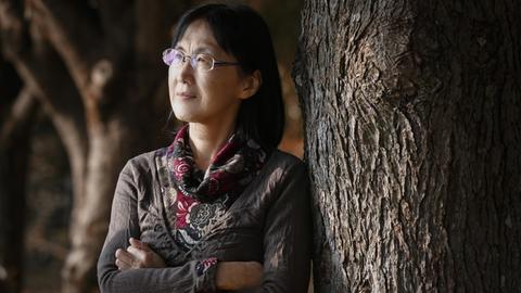 Noriko Kawakami mit verschränkten Armen an einen Baum gelehnt