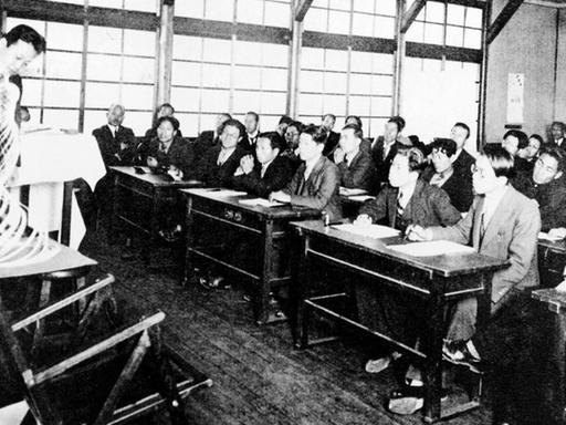 Das Bild zeigt japanische Studierende, die in einem Klassenraum eine Lehrer beobachten, der eine Vasen-Form vorführt.