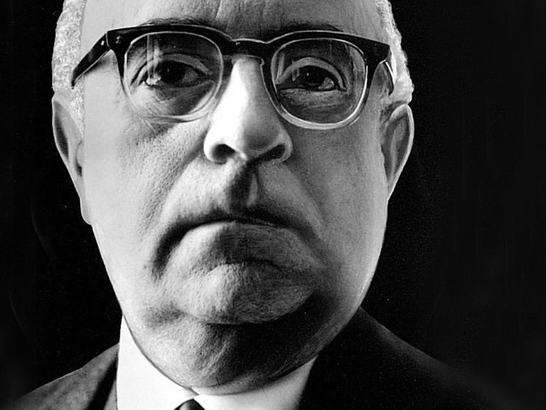 Ein Nahaufnahme von Theodor W. Adorno vor schwarzem Hintergrund. Er trägt eine Hornbrille und schaut aufmerksam dem Betrachter entgegen.