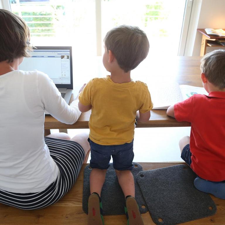 Eine Mutter sitzt mit ihren beiden kleinen Söhnen zuhause am Tisch, sie arbeitet am Laptop.