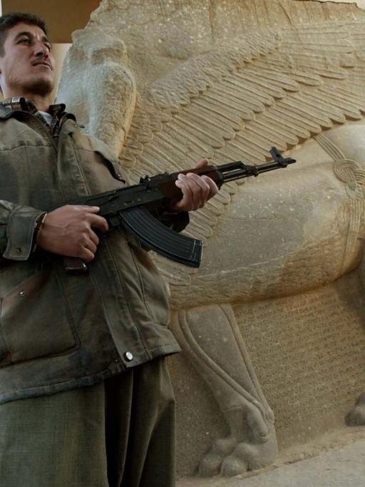 Ein mit einer Maschinenpistole bewaffneter Mann steht vor der steinernen Skulptur eines geflügelten Löwen.