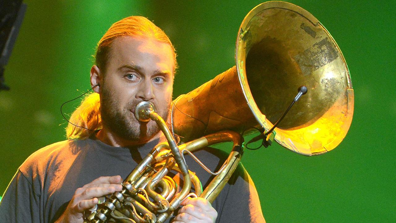 Ein Mann mit blondem Zopf steht Tuba spielend auf einer grün beleuchteten Bühne.