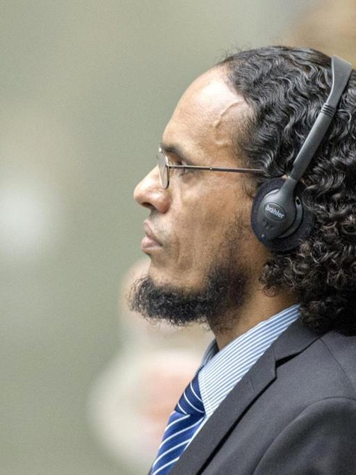 Das Bild zeigt Al Faqi Al Mahdi von der Seite auf der Anklagebank. Er trägt Kopfhörer und scheint jemandem aufmerksam zuzuhören. Er hat einen Anzug an und trägt eine Krawatte.
