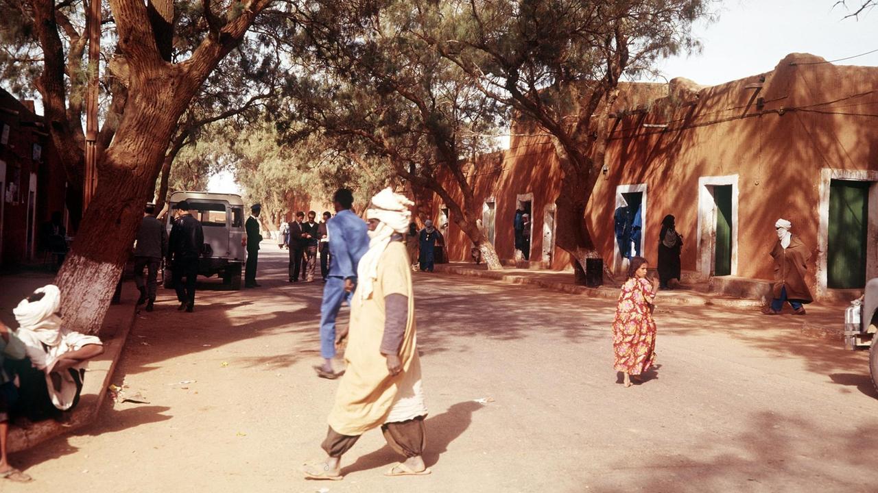 Blick in die Hauptstraße von Tamanrasset, dem Hauptort der Tuareg. Mehrere Menschen, u.a. mit Turbanen bekleidet, laufen über einen Platz mit einem großen Baum und einem Haus im Hintergrund.