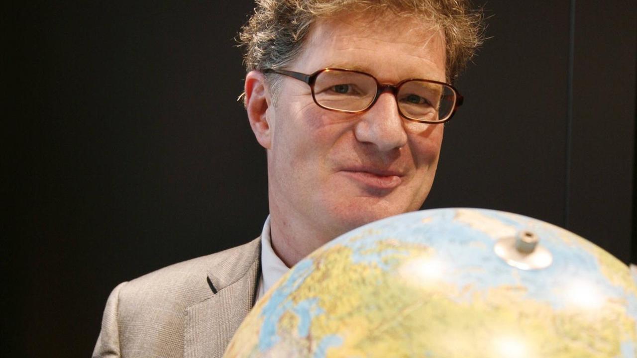 Ein Mann mit Brille hält einen Globus vor die Brust und schaut lächelnd darüber hinweg.