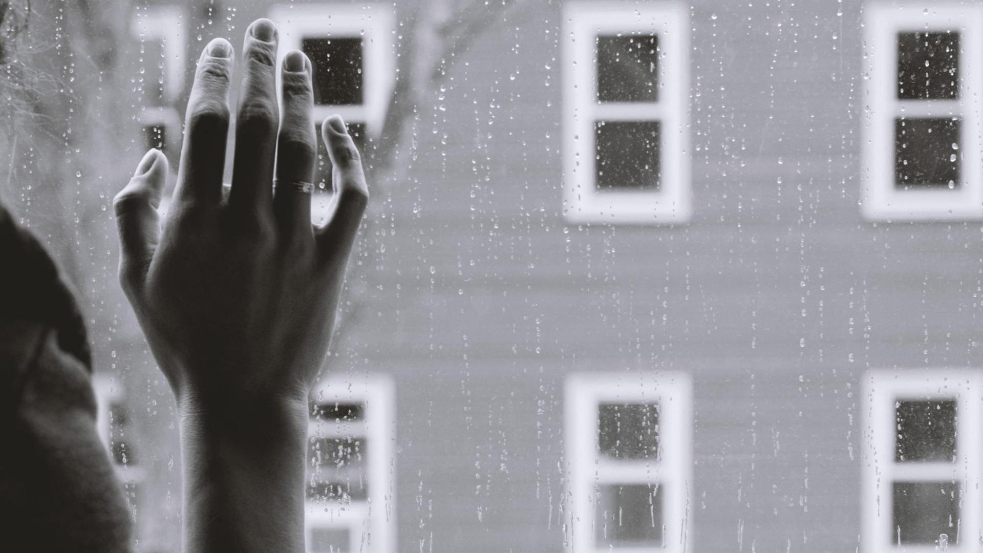 Schwarz-weiß-Bild einer Frauenhand an einer Fensterscheibe, draußen regnet es. Die Frau, deren Gesicht man nicht sieht, schaut offenbar auf die Straße.
