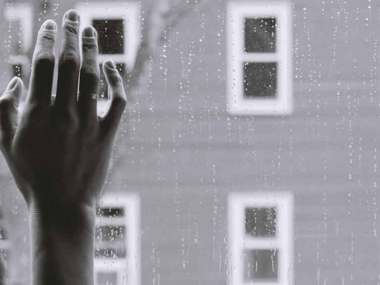 Schwarz-weiß-Bild einer Frauenhand an einer Fensterscheibe, draußen regnet es. Die Frau, deren Gesicht man nicht sieht, schaut offenbar auf die Straße.