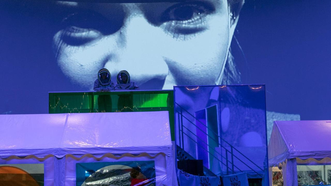 Auf einer Theaterbühne stehen im Vordergrund zwei blaue Plastikzelte. Dahinter stehen auf einer erhöhten Treppe zwei Darsteller in Weltraumanzügen und mit Astronautenhelmen, die sich dem Publikum zuwenden. Auf der Wand dahinter ist in Nahaufnahme das Gesicht der Schauspielerin Paula Kober projiziert.