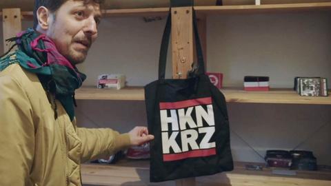 Pro7-Reporter Thilo Mischke steht vor einem Regal und zeigt eine schwarze Stofftasche, die mit der Abkürzung "HKN KRZ" für "Hakenkreuz" bedruckt ist.