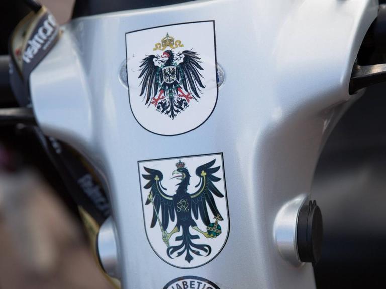 Mehrere Wappen als Statement für eine Monarchie nach Vorbild des Deutschen Reichs kleben auf einem kleinen Wagen für Menschen mit Gehproblemen.
