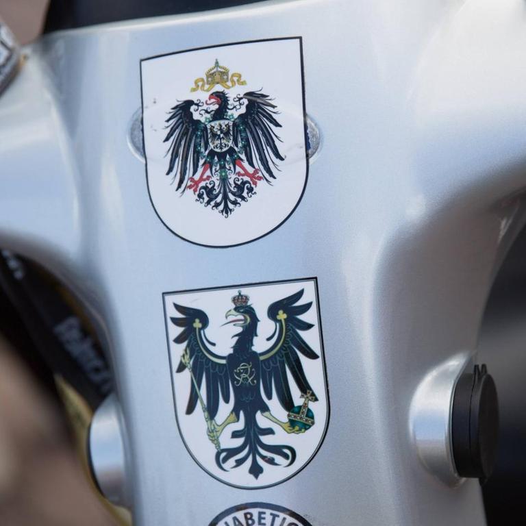 Mehrere Wappen als Statement für eine Monarchie nach Vorbild des Deutschen Reichs kleben auf einem kleinen Wagen für Menschen mit Gehproblemen.