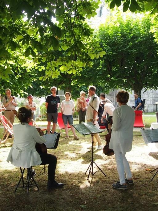 Drei Musikerinnen und eine Gruppe von Menschen haben sich im Schatten der Bäume versammelt