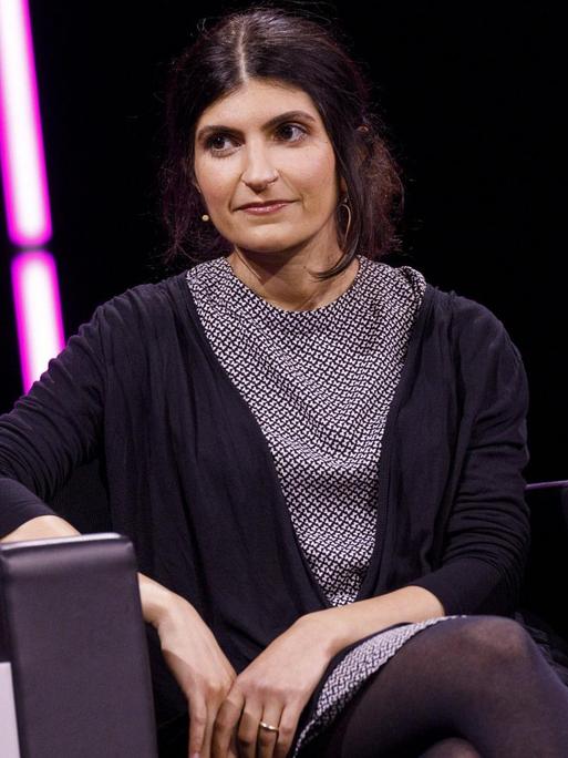 Shida Bazyar auf einem Sessel auf einer Bühne, im Hintergrund sind rosa Neonröhren zu sehen