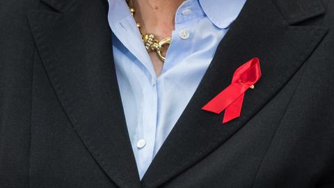 Eine Person trägt die rote Schleife, das weltweite Symbol für Solidarität mit Hiv-Infizierten und Aids-Kranken, am Revers eines schwaren Blazers und darunter eine hellblaue Bluse.