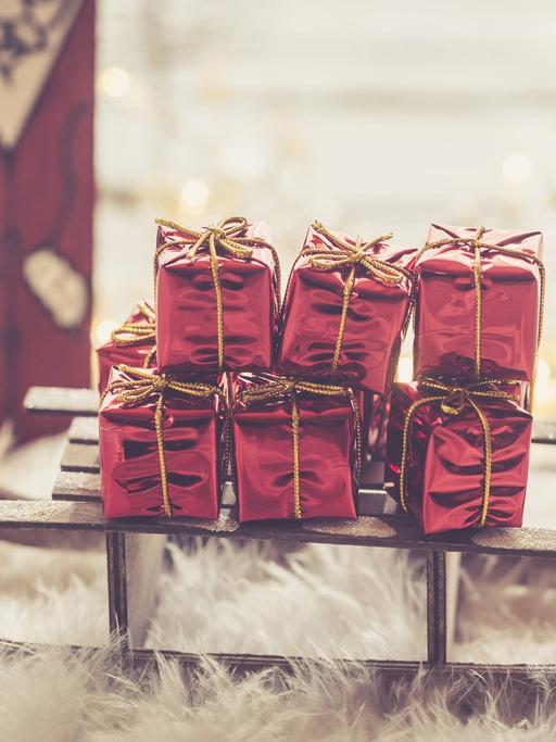Eine kleine Weihnachtsmannfigur steht hinter einem Holzschlitten, der mit roten Geschenken beladen ist.