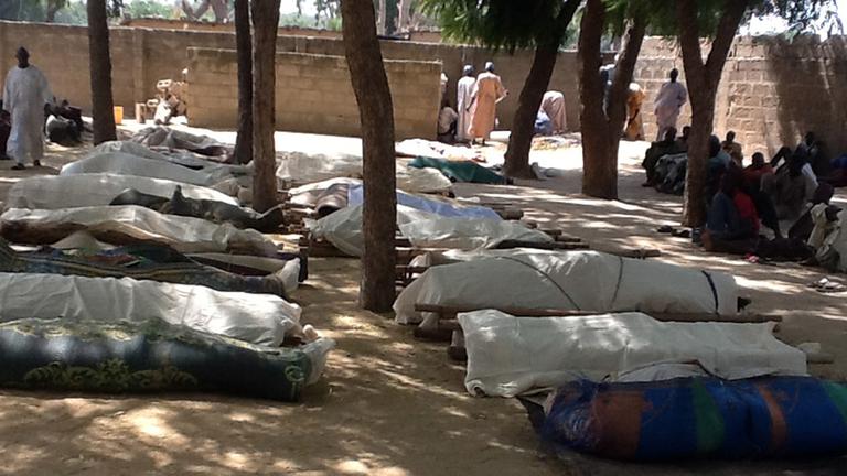 Im Ort Konduga liegen mehrere Leichensäcke aus. Menschen sitzen in der Nähe.