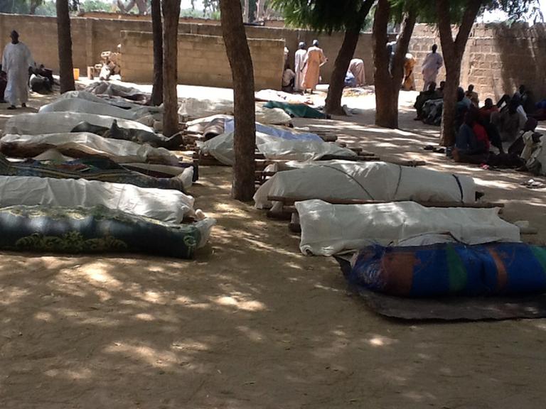 Im Ort Konduga liegen mehrere Leichensäcke aus. Menschen sitzen in der Nähe.