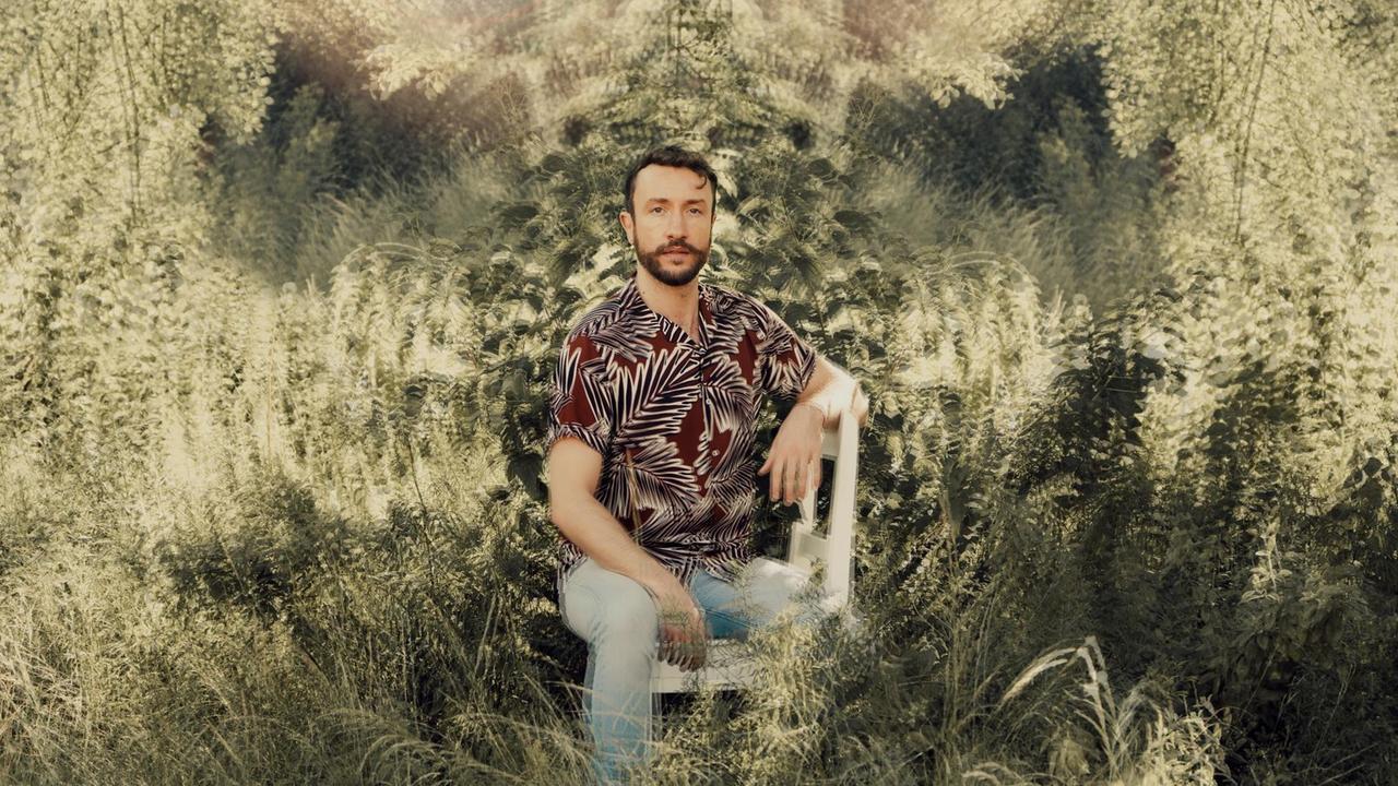 Ein Mann in buntem Hemd sitzt in der Natur. Das Bild ist zum Rand hin leicht verfremdet, sodass es wirkt, als säße er vor einem Tamtam.