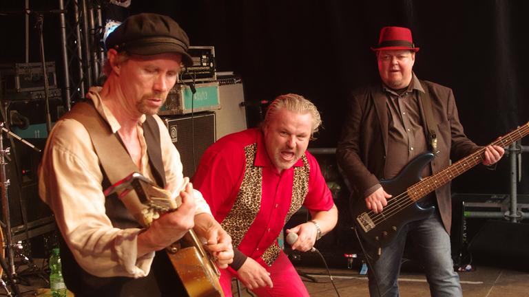 Drei Männer stehen musizierend auf einer Bühne: einer spielt Gitarre, der Mann in der Mitte hält ein Mikrofon in der Hand, hinter ihm steht ein Bassist.