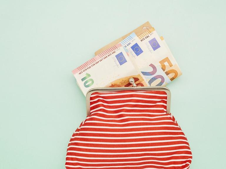 Eine rotweiss gestreifte Geldbörse liegt auf einem mintfarbenen Untergrund. Daraus schauen einige Euronoten.