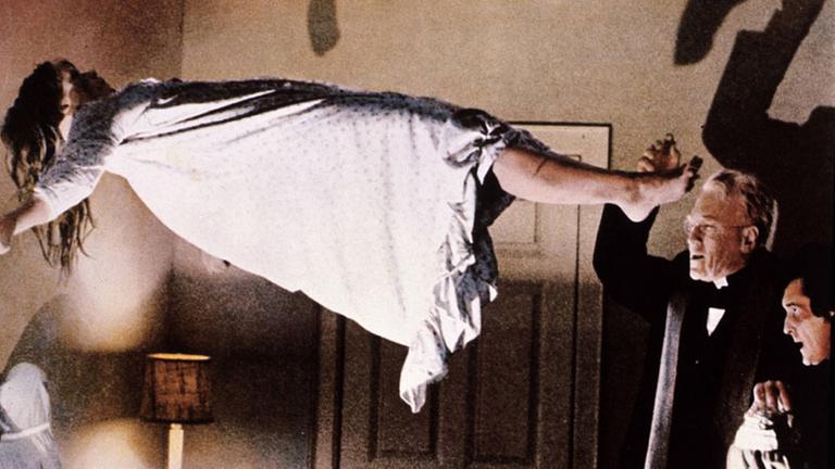 Szene aus dem Horrorfilm "Der Exorzist". Ein Mädchen im Nachthemd schwebt in einem Zimmer in der Luft, ein Priester macht beschwörende Gesten