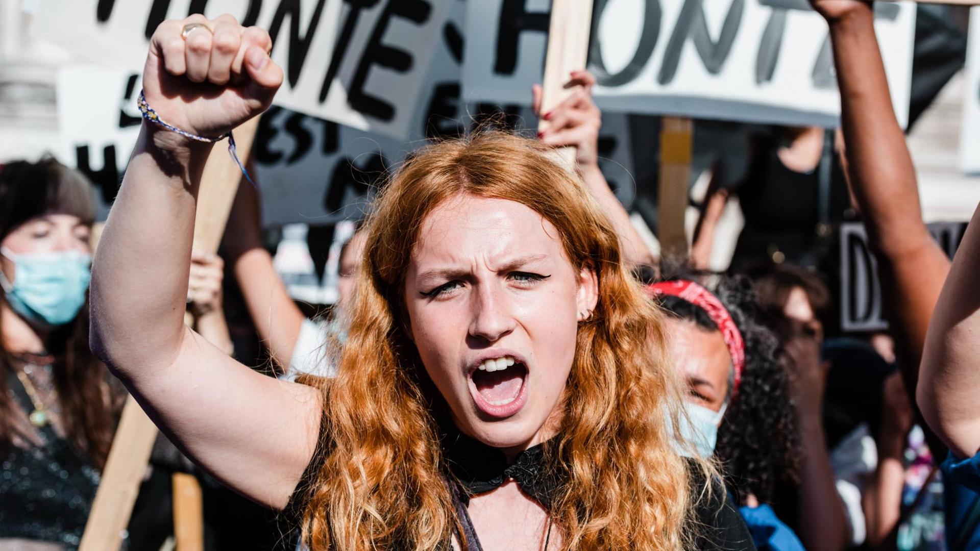 Frauendemonstration in Paris: Im Mittelpunkt des Bildes eine junge Frau mit rotblonden langen Haaren, die eine Faust in die Höhe gereckt hat. Im Hintergrund weitere in die Höhe gereckte Fäuste und Plakate.